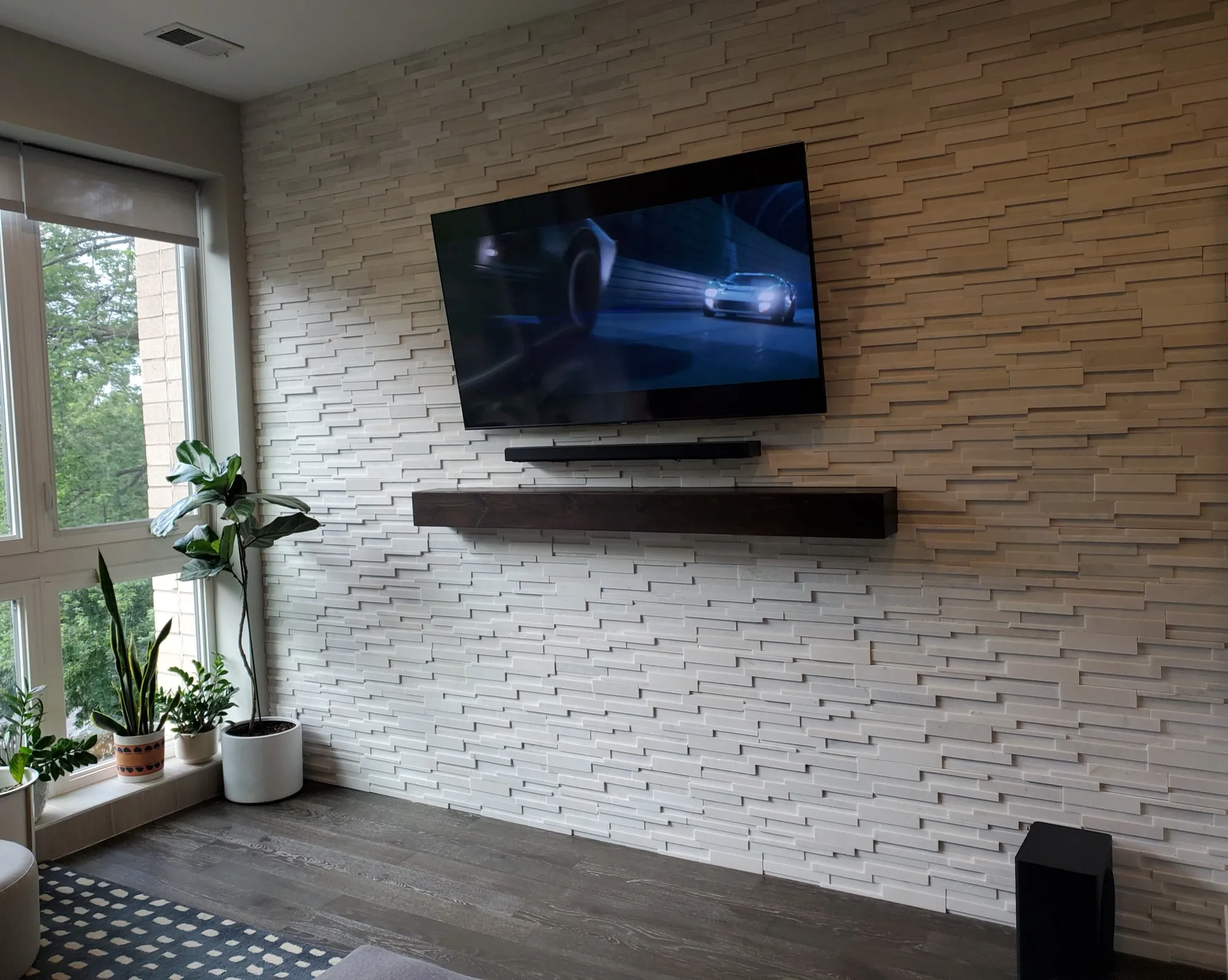 Large television mounted flush on a stacked stone accent wall above a floating walnut shelf, with a soundbar below the screen.